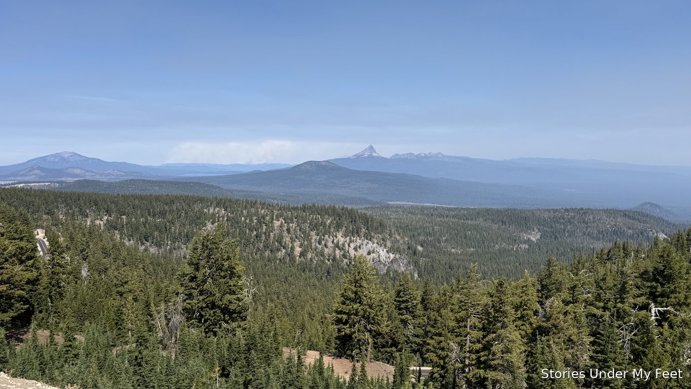 A sweep of evergreen forest and a distant snow-capped peak seen from the rim road